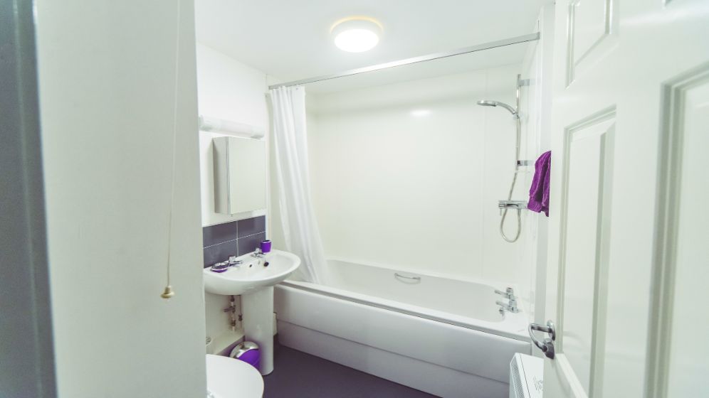 A photo taken from the bathroom door. There is a bath with a shower over it. There's a sink with a mirrored unit above it, as well as a toilet.